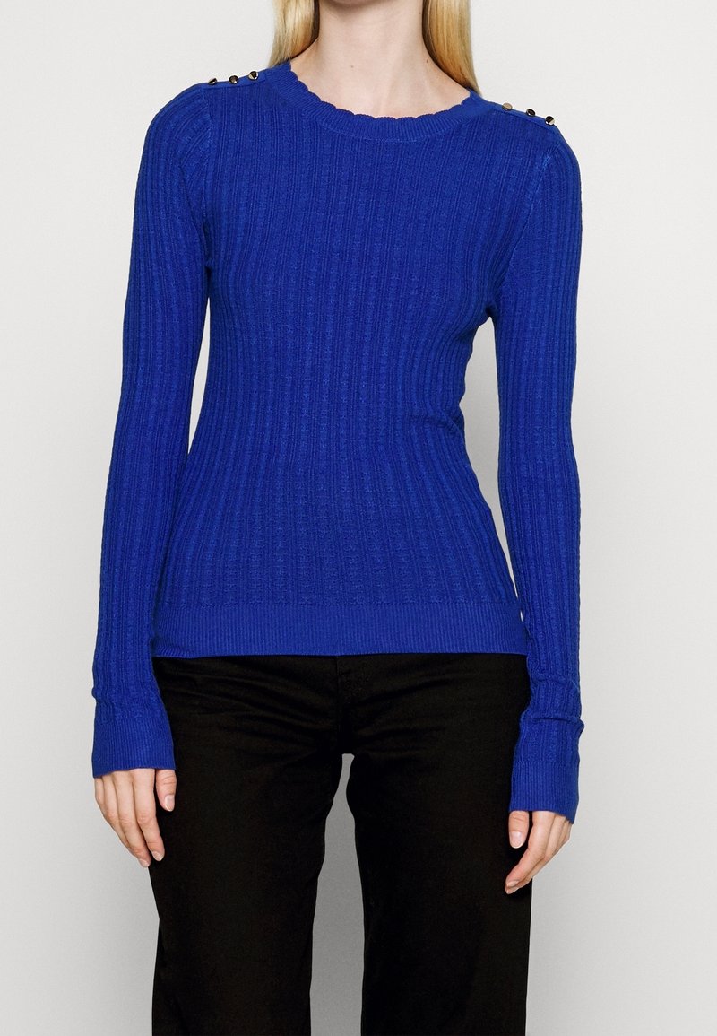 Femme portant un pull côtelé bleu roi à manches longues avec des détails de boutons sur les épaules et un pantalon noir sur un fond uni.