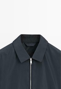 Mørk navy jakke med spids krave og sølvfarvet lynlås foran, vist fra brystet og op mod en hvid baggrund.