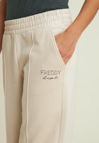 Pantalones de chándal beige claro con cintura elástica, bolsillo lateral y texto "FREDDY dept" en piedras decorativas en la pierna izquierda.