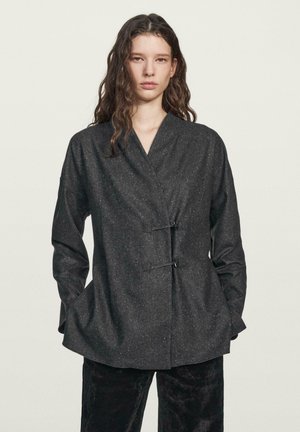 Black textured jacket with a wrap design, featuring a v-neckline and two decorative clasps. Long sleeves and side pockets complete the look.