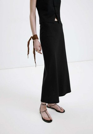 Femme en haut noir sans manches et jupe avec un bracelet en tissu marron au poignet, portant des sandales plates noires à brides sur un sol blanc.