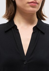 Žena u crnoj košulji s ovratnikom i dubokim V-izrezom s malim gumbom, vidi se od vrata do gornjeg dijela prsa.