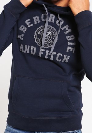 Person wearing navy blue Abercrombie and Fitch hoodie with front pocket and grey drawstrings, adjusting the hood.