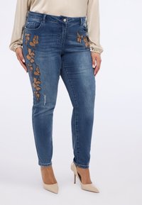 Ciemnoniebieskie jeansy z denimu z kwiatowym haftem w metalicznym odcieniu brązu, dopasowany krój i lekko przetarte detale na prawej udzie.