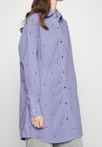 Lys lilla skjorte med knap til op knapning, der har et subtilt stribet mønster og broderede motiver. Har krave, lange ærmer og sorte knapper.