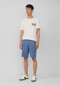 Lichtbeige T-shirt met marineblauwe grafische print, gecombineerd met blauwe shorts. Witte sneakers met bruine accenten. Eenvoudig ontwerp, korte mouwen.