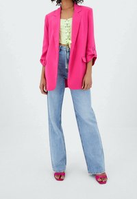 Mujer vestida con una chaqueta fluorescente rosa, top corto de encaje verde claro, jeans de mezclilla azul claro y tacones abiertos rosa a juego, de pie sobre un fondo blanco.