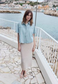 Vrouw met halflang haar, gekleed in een licht denim overhemd, een beige gebreide rok en sandalen, staand bij een zeezichtleuning op een betegeld terras.