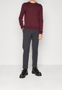 Homme portant un pull bordeaux à manches longues, un pantalon gris foncé sur mesure, et des chaussures noires à lacets épaisses, debout avec une main dans la poche.