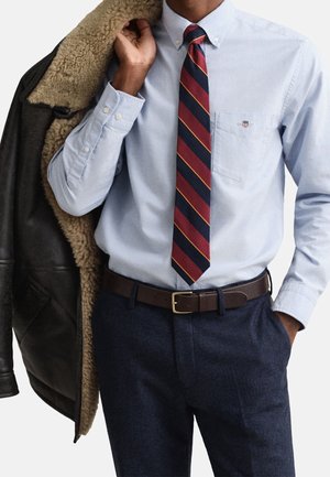 Man wearing light blue button-down shirt, striped red and navy tie, navy pants, brown belt, holding dark brown shearling-lined leather jacket.