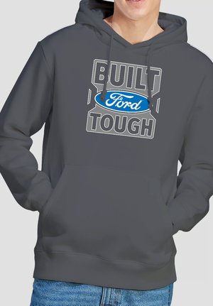 Person trägt einen dunkelgrauen Hoodie mit dem Aufdruck "BUILT FORD TOUGH" und blauem Ford-Logo, die Hände in der vorderen Tasche, kombiniert mit blauen Jeans.