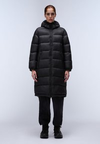 Zwarte pufferjas met capuchon, met een horizontaal gestikt patroon, voorrits en zijzakken; gedragen over zwarte broek en laarzen.