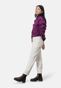 Fioletowa koszula z długim rękawem z dwoma przednimi kieszeniami, zestawiona z białymi prążkowanymi spodniami i czarnymi botkami za kostkę z grubą podeszwą.