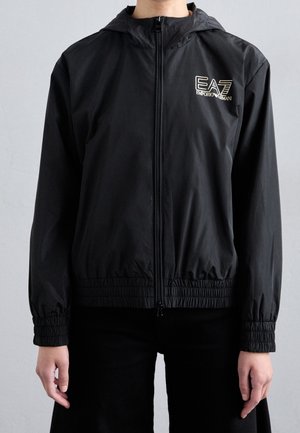 Osoba nosząca czarną kurtkę z kapturem na zamek błyskawiczny, z elastycznymi mankietami i logo EA7 Emporio Armani na piersi, stojąca na jednolitym tle.