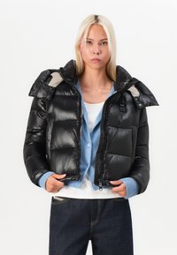 Schwarze Pufferjacke mit glänzender Textur, mit Reißverschluss vorne, großem Kragen und blauen Akzenten darunter, kombiniert mit einer hellblauen Strickjacke und dunklen Jeans.