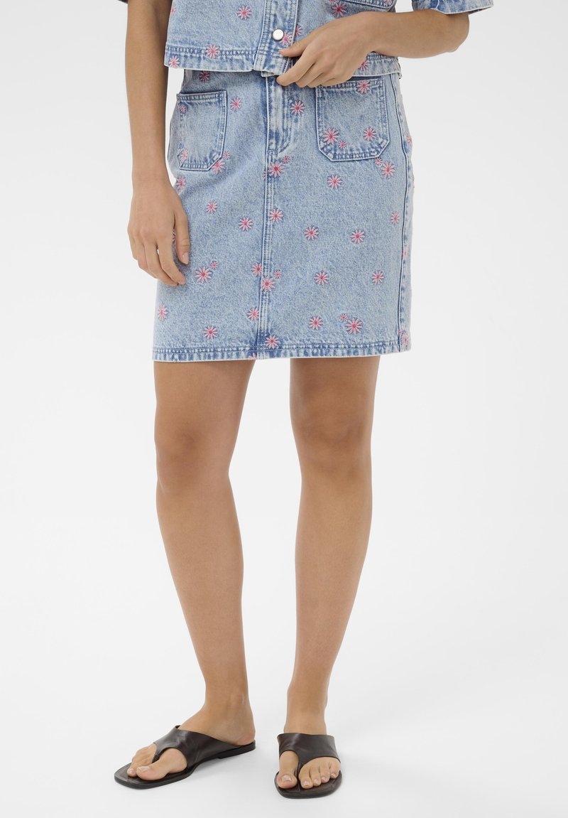 Jupe en denim avec broderie florale rose, poches avant, portée avec un crop top assorti et des sandales plates noires à bout ouvert sur jambes nues.