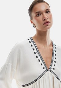 Blusa bianca con scollatura a V caratterizzata da ricami floreali neri e bordi. Tessuto testurizzato con maniche lunghe e dettagli arricciati.