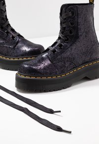 Paire de bottes en cuir noir craquelé à lacets, semelles épaisses et coutures jaunes, une botte montrée en gros plan avec des lacets noirs desserrés à l'avant.
