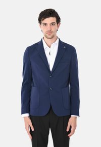 Blazer navy realizzato in tessuto liscio con due tasche frontali, chiusura a un bottone e un piccolo accento con logo sulla patta. Camicia bianca sotto.