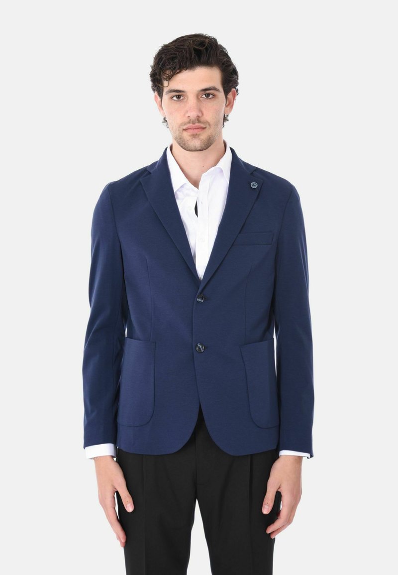 Blazer navy realizzato in tessuto liscio con due tasche frontali, chiusura a un bottone e un piccolo accento con logo sulla patta. Camicia bianca sotto.