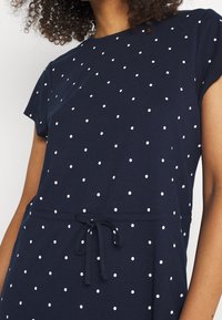 Robe bleue marine en tissu doux, ornée de pois blancs, avec manches courtes et taille à cordon de serrage pour un ajustement personnalisé.