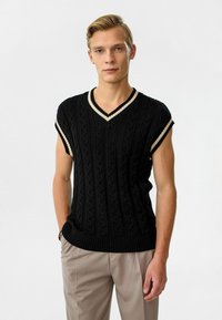 Zwarte kabelgebreide mouwloze trui met een V-hals en crèmekleurige afwerking op de kraag en armsgaten, gecombineerd met lichtbeige broeken.