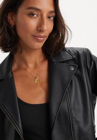 Musta nahkatakki, jossa on lovetettu kaulus, hopeiset yksityiskohdat ja vinoketju. Takki on päällä mustan ribbipintaisen topin, jossa on kultaketju.