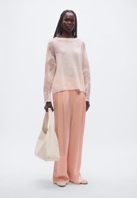 Leichter, strukturierter Pullover in weichen Rosatönen, kombiniert mit fließenden, hoch taillierten Hosen in Pastellrosa. Hält eine cremefarbene, weich gearbeitete Handtasche.