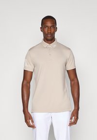Beige polo skjorte med krage og tre knapper, med et teksturert mønster på venstre erme. Laget av et glatt, elastisk stoff.