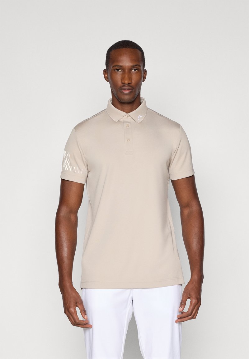 Beige polo skjorte med krage og tre knapper, med et teksturert mønster på venstre erme. Laget av et glatt, elastisk stoff.