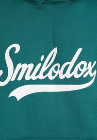 Sudadera color verde azulado con un gran logotipo en blanco de letra manuscrita "Smilodox" en el pecho, hecha de un tejido suave con una textura lisa.