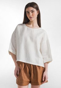 Ung kvinde med langt brunt hår iført løs hvid bluse med albuelange ærmer og højtaljede brune shorts mod hvid baggrund.