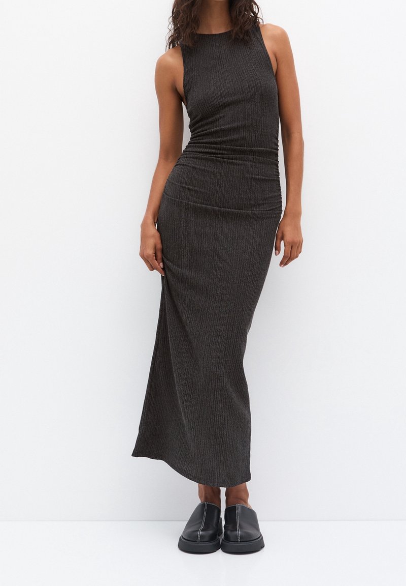 Femme portant une robe maxi sans manches côtelée gris foncé et des chaussures noires sans lacets, debout devant un fond blanc.