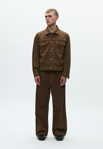 Καφέ τζιν μπουφάν με δύο τσέπες στο στήθος και κουμπιά, σετ με παντελόνι φαρδύ γκάζι του ίδιου χρώματος. Ομαλή υφή, λιτός σχεδιασμός. Μαύρα παπούτσια.