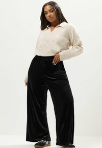 Beige stickad tröja med krage som matchas med svarta vida sammetbyxor. Outfiten kompletteras av svarta slip-on skor med vitt mönster.