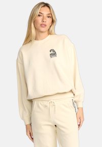 Felpa crema con maniche lunghe e polsini a costine; presenta un piccolo logo grafico nero sul petto. Abbinata a pantaloni della tuta coordinati.