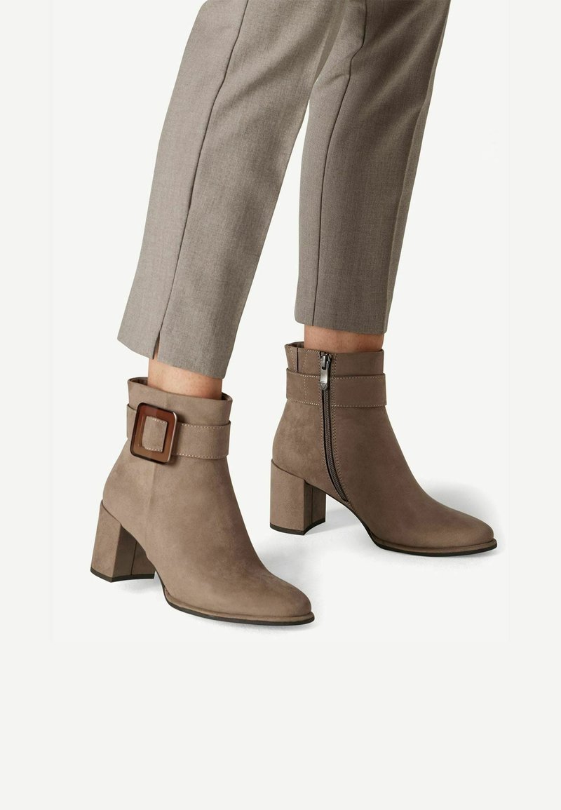 Bottes en daim taupe à hauteur de cheville avec talons blocs, fermetures éclair latérales et grandes boucles décoratives, portées avec un pantalon beige court.