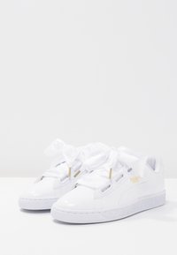 Baskets blanches Puma Basket avec des lacets larges en ruban satin et des accents dorés sur un fond blanc immaculé.