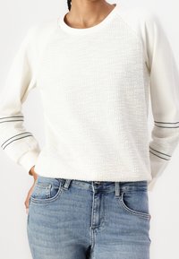 Valkoinen teksturoitu collegepaita, jossa on kiinteä yoke ja raidoitukset hihoissa, yhdistettynä keskipestyihin sinisiin farkkuihin.