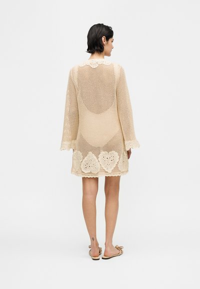 Γυναίκα με κοντά σκοτεινά μαλλιά που φοράει μπεζ πλεκτό φόρεμα με σχέδια καρδιές κοντά στο τελείωμα, όρθια ξυπόλητη σε λευκό φόντο, με την πλάτη προς τον θεατή.