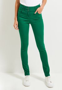 Jeans skinny verdi realizzati in tessuto elasticizzato, con vita alta, due tasche frontali e chiusura a due bottoni, abbinati a sneaker bianche.