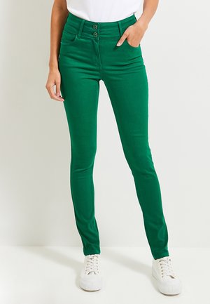 Jeans skinny verts en tissu extensible, avec une taille haute, deux poches avant et une fermeture à deux boutons, associés à des baskets blanches.