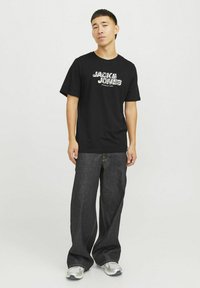 Sort t-shirt med "JACK & JONES" logo i et kontrasterende mønster. Kombineret med løstsiddende mørke denim jeans og grå sneakers.