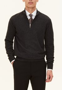 Dunkelgrauer Strickpullover mit einem Reißverschlusskragen, Slim Fit und gerippten Bündchen. Über einem weißen Hemd getragen und mit schwarzen Hosen kombiniert.