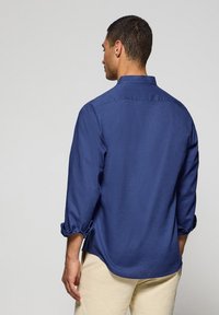 Granatowa koszula z długim rękawem, z podwijanymi rękawami, z kołnierzykiem zapinanym na guziki oraz detalem na plecach. Wykonana z lekkiego materiału o gładkiej fakturze.