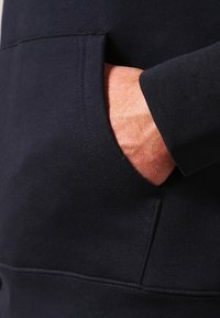 Marineblaue Sweatshirt-Stoff mit Kängurutasche und gerippten Bündchen. Nahaufnahme zeigt eine Hand, die in der Tasche ruht, und hebt die Textur hervor.