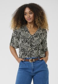 Blouse noire à manches courtes avec motif de feuilles blanches, décolleté en V et accents beige clair. Assortie à un pantalon en denim bleu taille haute et une ceinture marron.