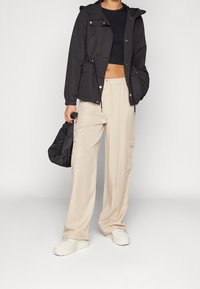 Giacca nera con cappuccio e coulisse regolabili, abbinata a pantaloni cargo beige a gamba larga con tasche laterali e sneaker bianche con plateau.