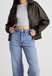Personne portant une veste en cuir noire par-dessus un top gris court et un jean bleu taille haute, tenant la veste ouverte avec les deux mains.