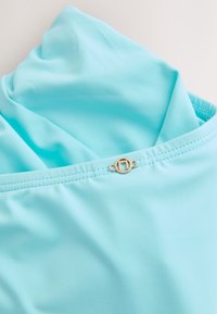 Gros plan d’un tissu bleu clair avec des coutures détaillées et une petite boucle métallique ronde avec un motif gravé.